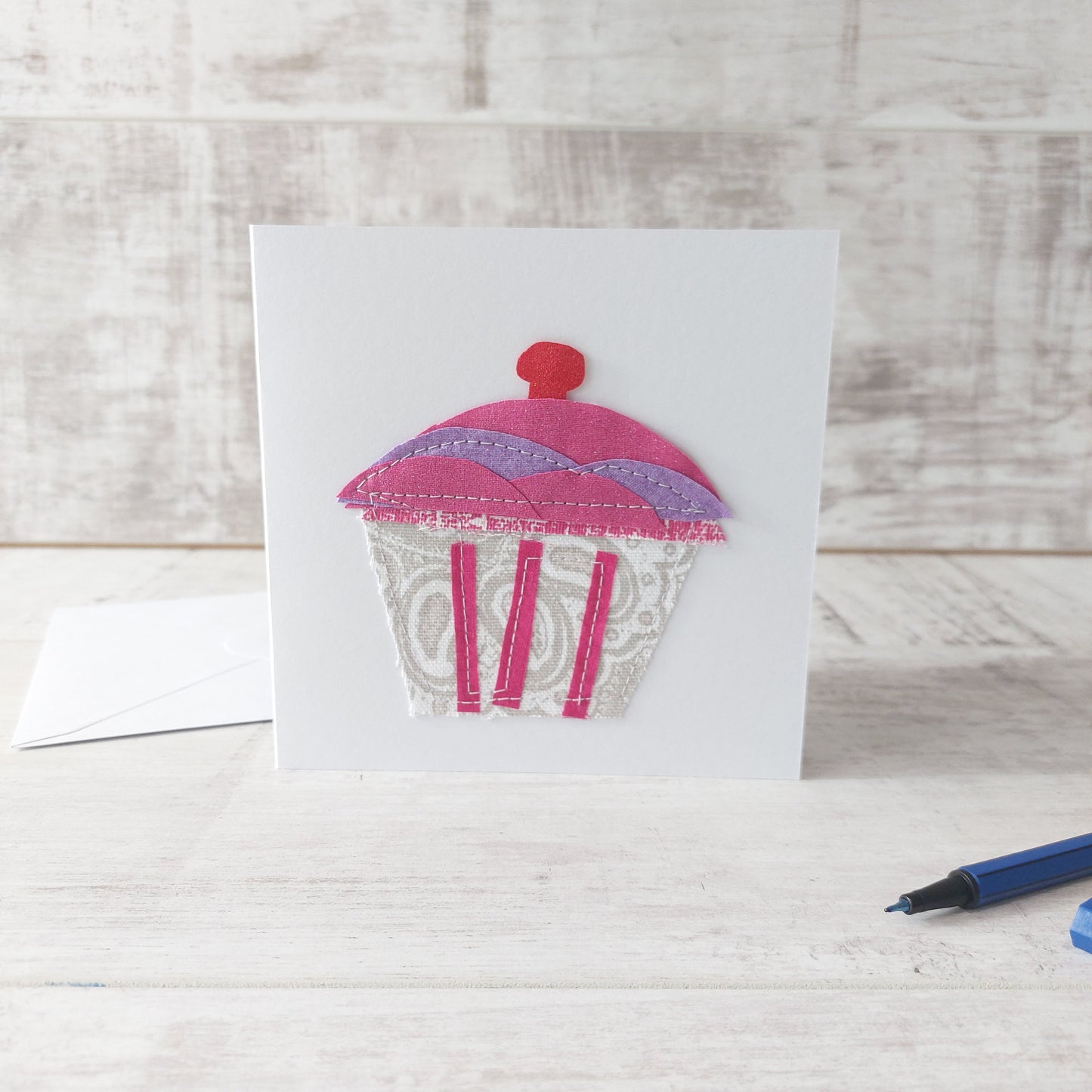 Greetings card featuring a fabric cupcake applique with a cherry on top, pictured with an envelope and pen, by Olganna.