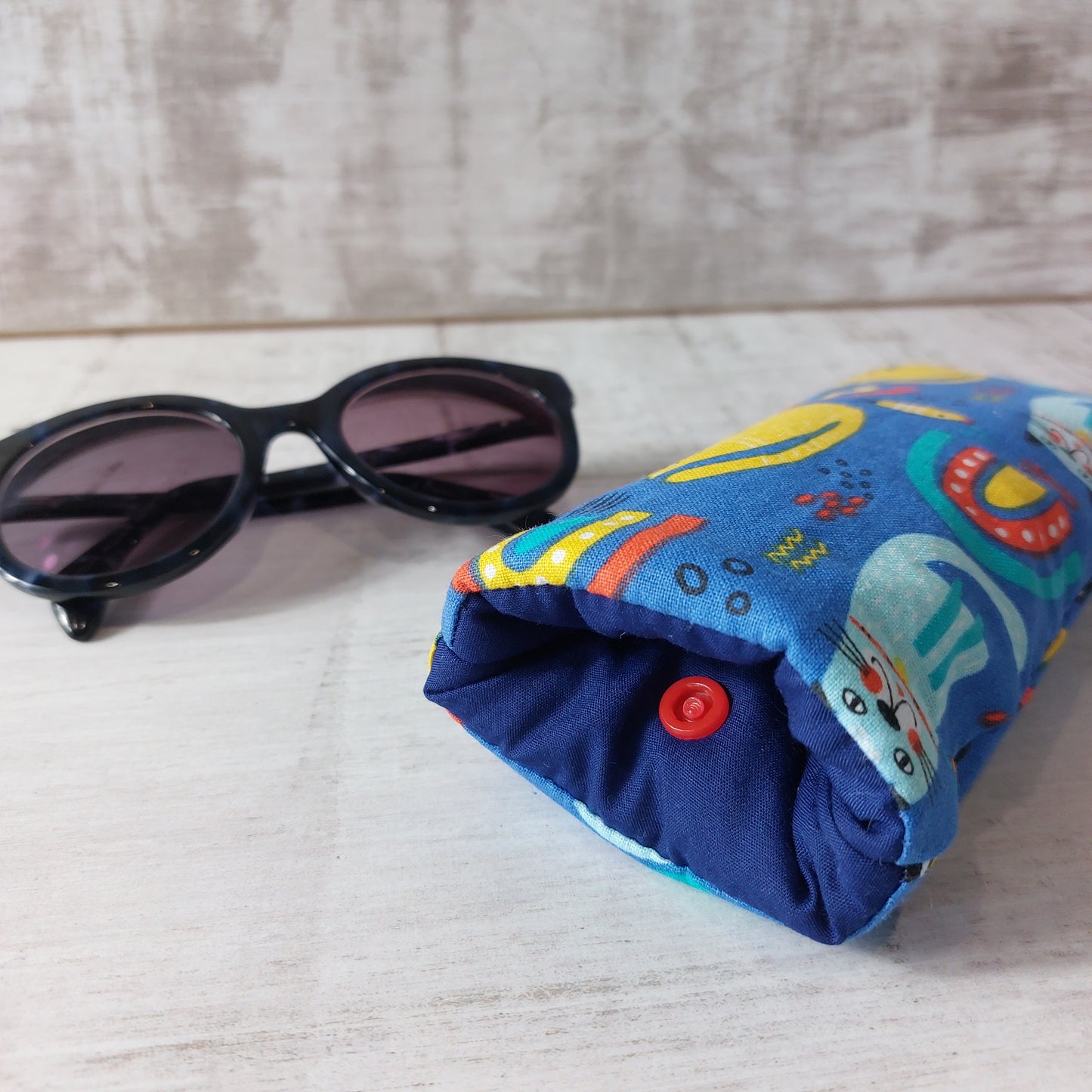 Close-up of blue rainbow and cats printed glasses case showing press stud fastening, pictured with sunglasses.