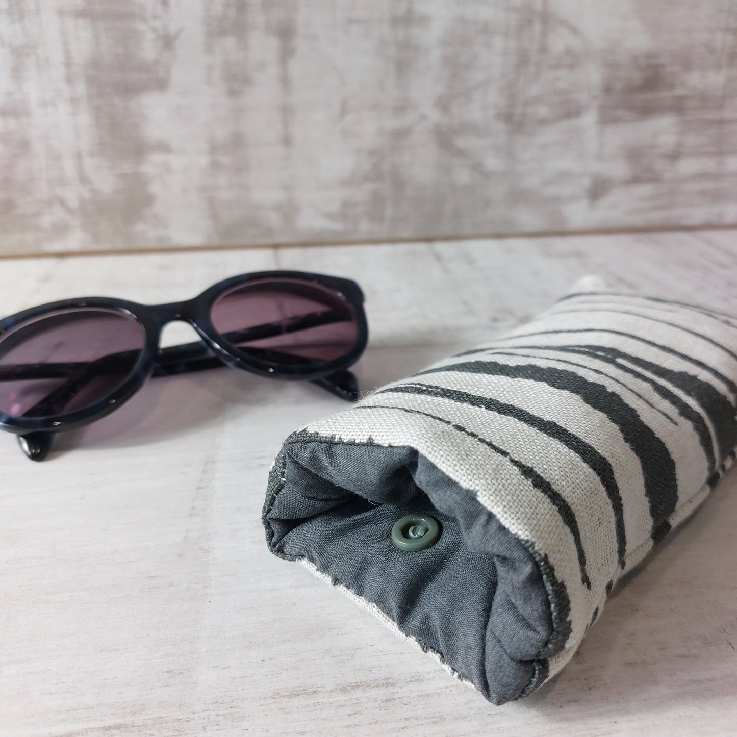 Closeup of neutral stripe linen based fabric sunglasses case with snap fastening, pictured with sunglasses.