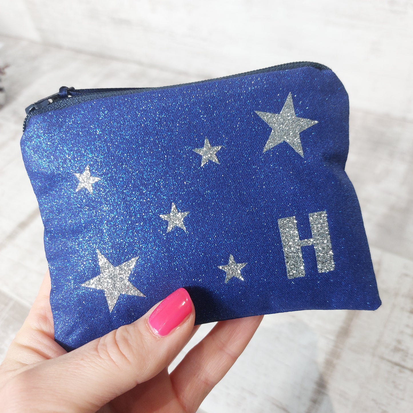 A hand holding a sparkly blue zip purse with silver glitter stars and personalised with a silver glitter letter 'H', by Olganna.