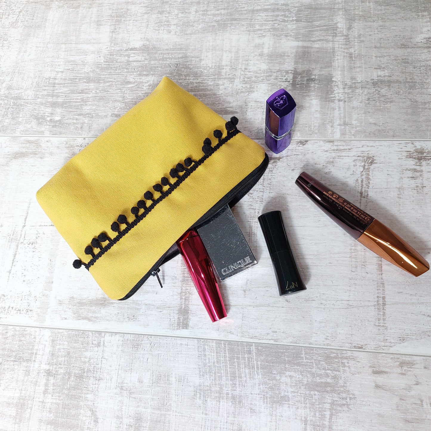 Mustard canvas makeup bag with black pom poms, pictured with beauty essentials.