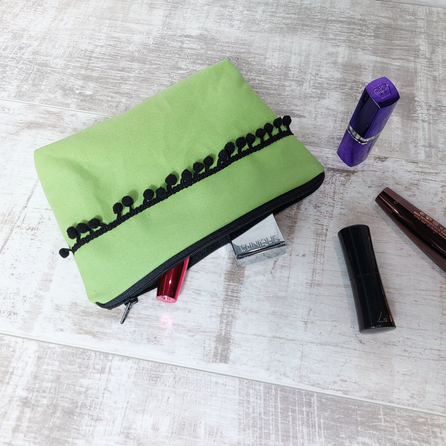 Lime green canvas makeup bag with black pom poms, pictured with beauty essentials.