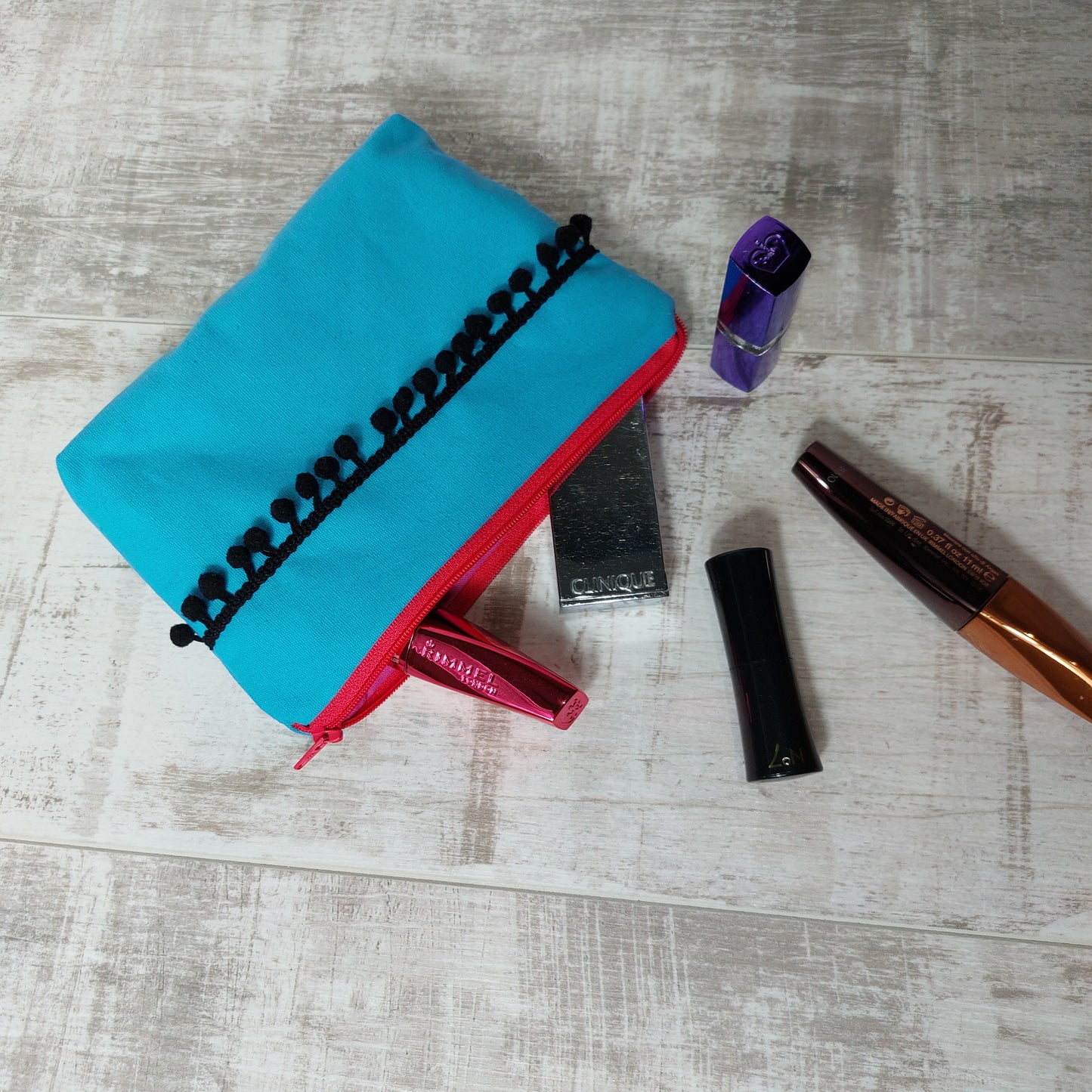 Turquoise canvas makeup bag with black pom poms, pictured with beauty essentials.