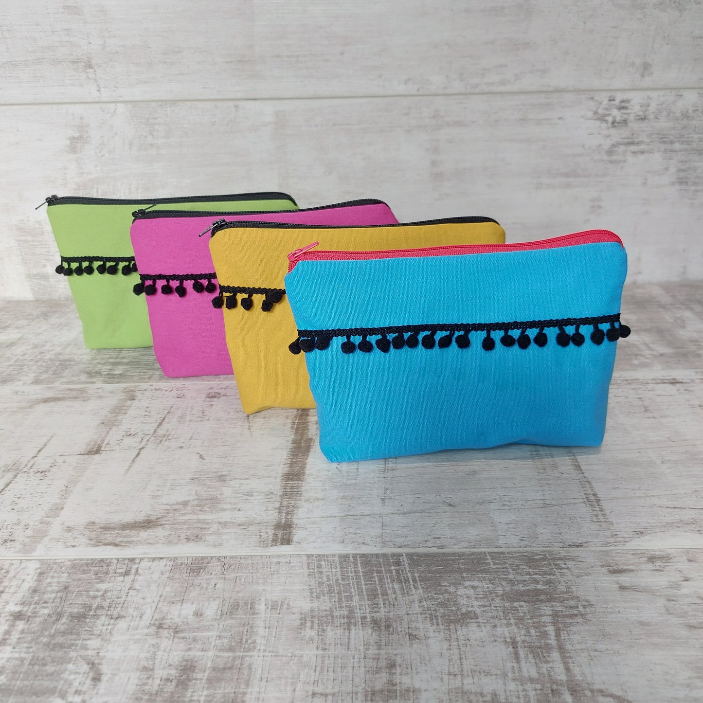 Four canvas makeup bags with black pom poms, in turquoise, mustard, hot pink and lime green, lined up.