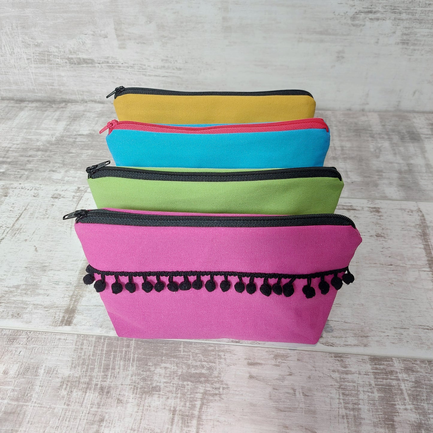 Four canvas makeup bags, lined up, in hot pink, lime green, turquoise and mustard, with black pom poms.