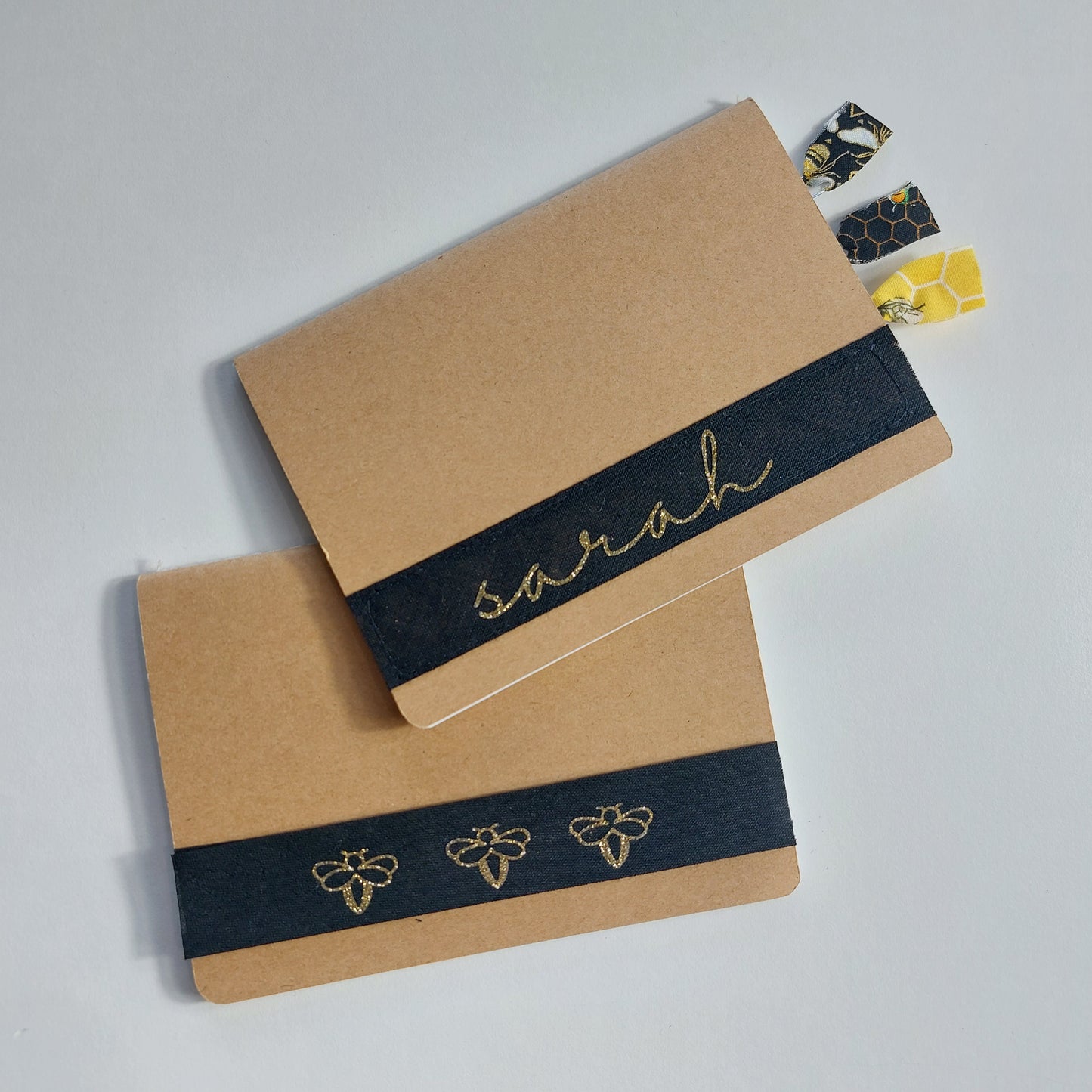 Two notepads with a kraft card cover with gold glitter print on a stitched black band background, one personalised with 'Sarah' and one with a row of three bees.