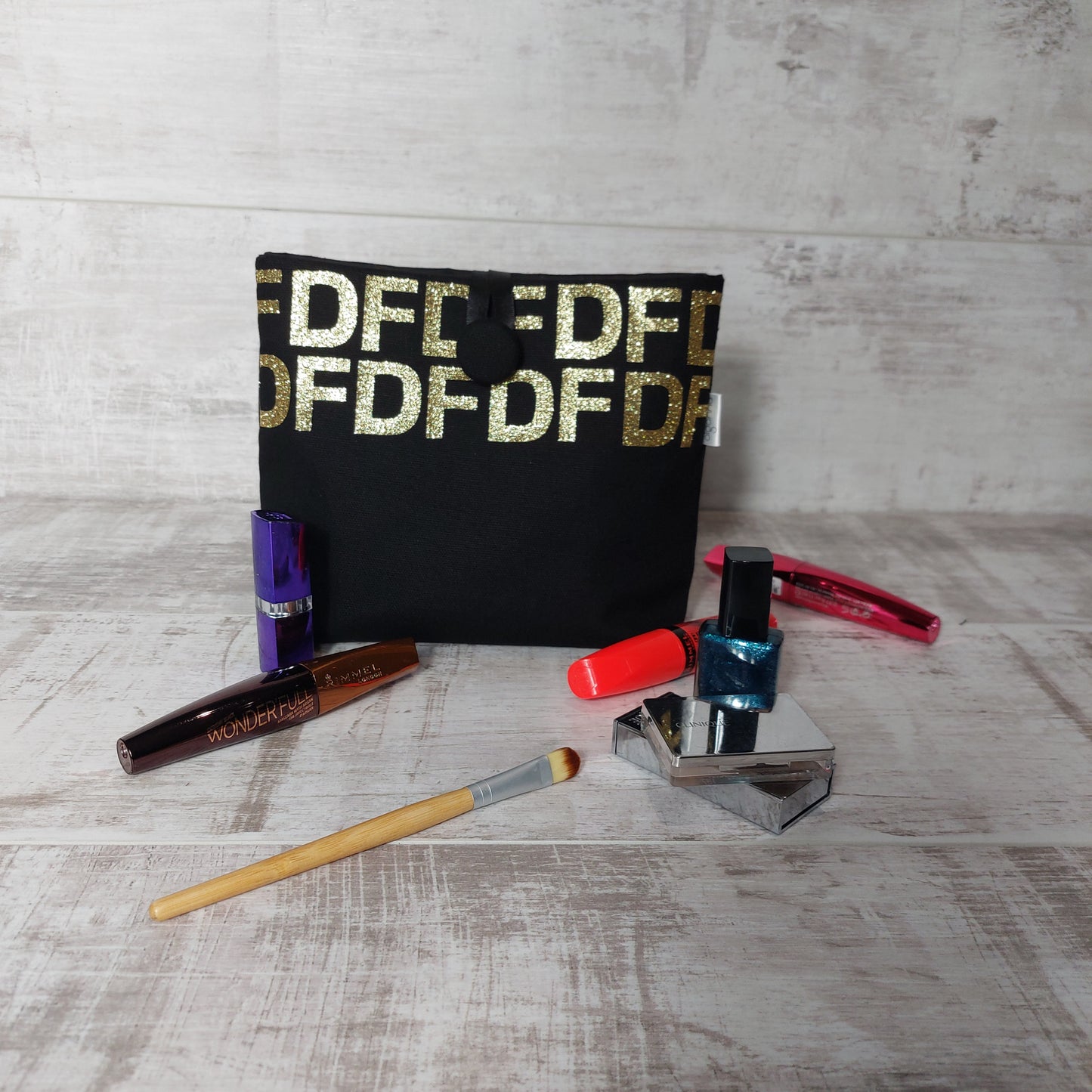 A button pouch in black canvas fabric personalised with gold glitter initials 'DF' in repeating print, standing upright with assorted makeup products in the foreground.