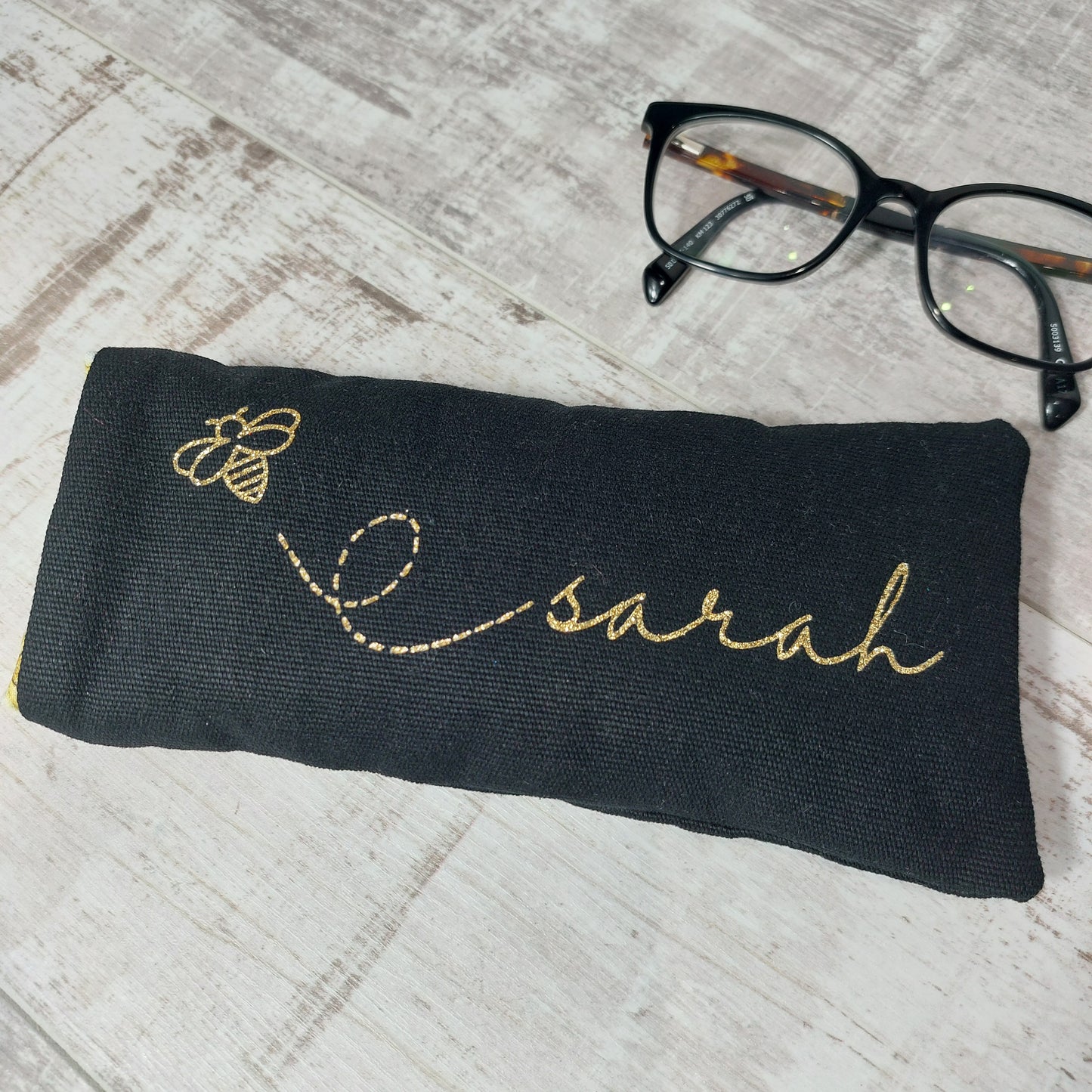 Black soft padded glasses case with gold glitter bee and personalised with the name 'Sarah' in gold, pictured with glasses.
