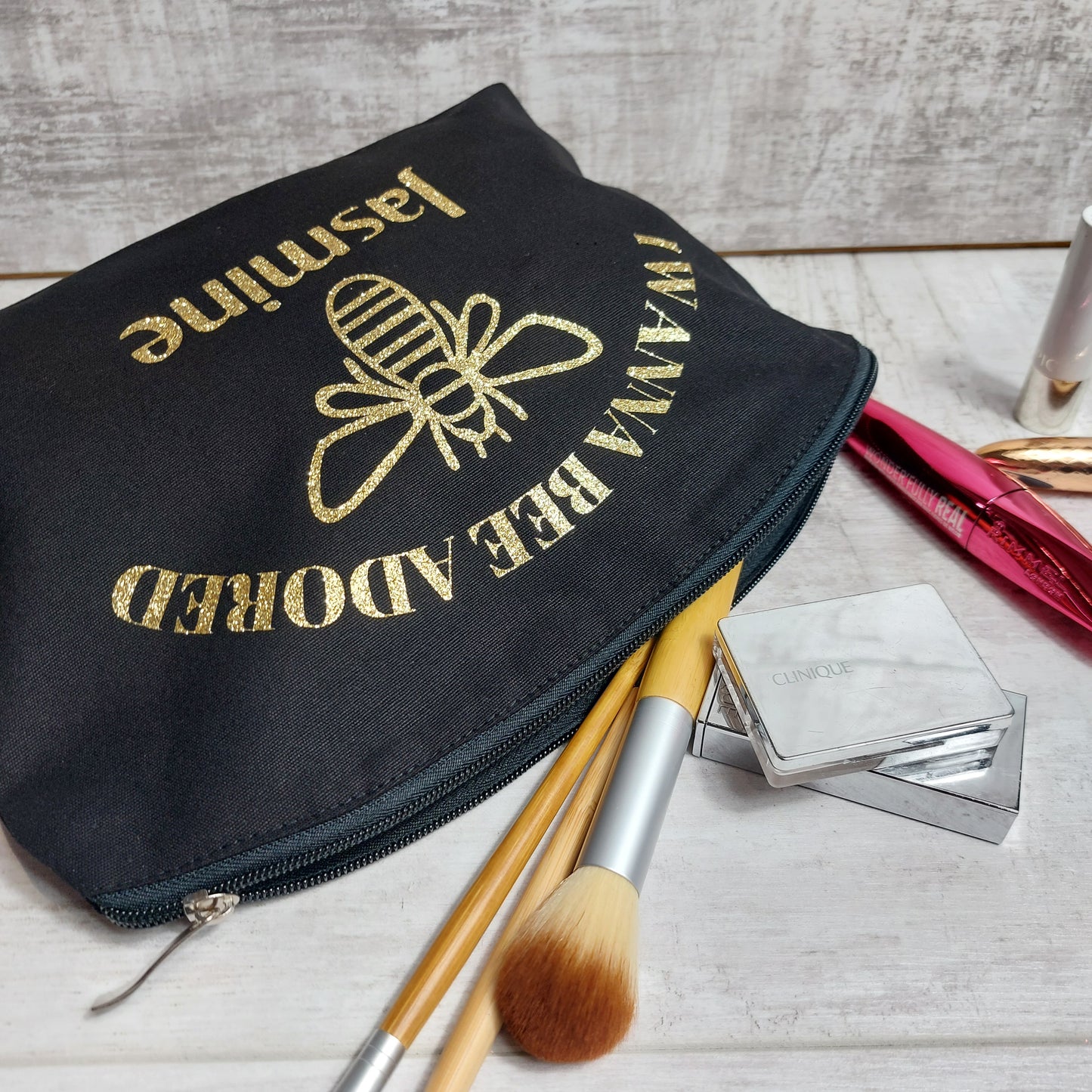 Large rounded top makeup bag in black with gold glitter bee under text 'I WANNA BEE ADORED' and personalised with the name 'Jasmine' also in gold glitter, pictured lying down and unzipped to reveal makeup and brushes.