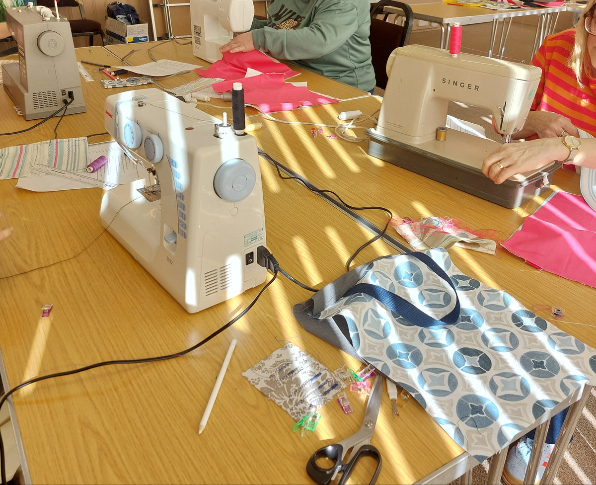 Person using a sewing machine at a table with various items including a patterned bag and scissors.