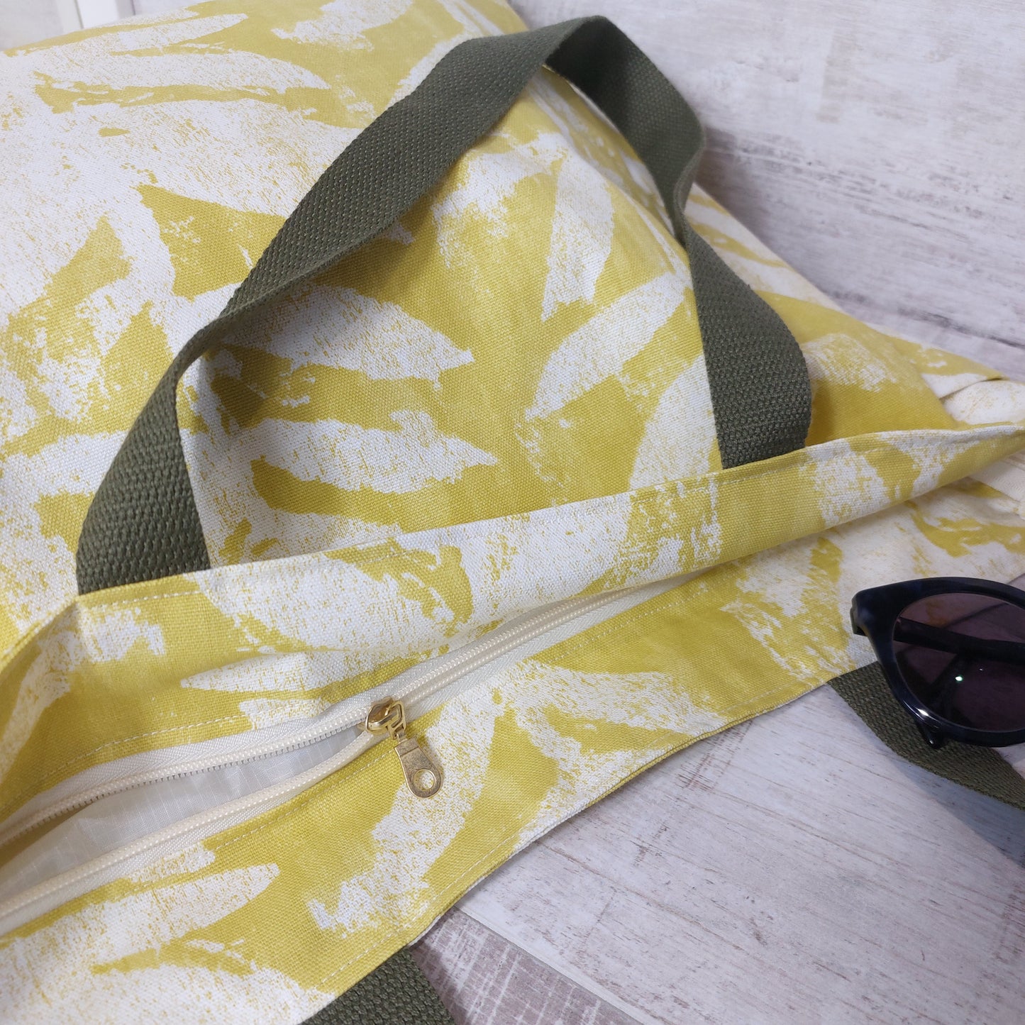 Top opening of bright citrus yellow leafy print beach bag, with deep-set zip closure, pictured with sunglasses.