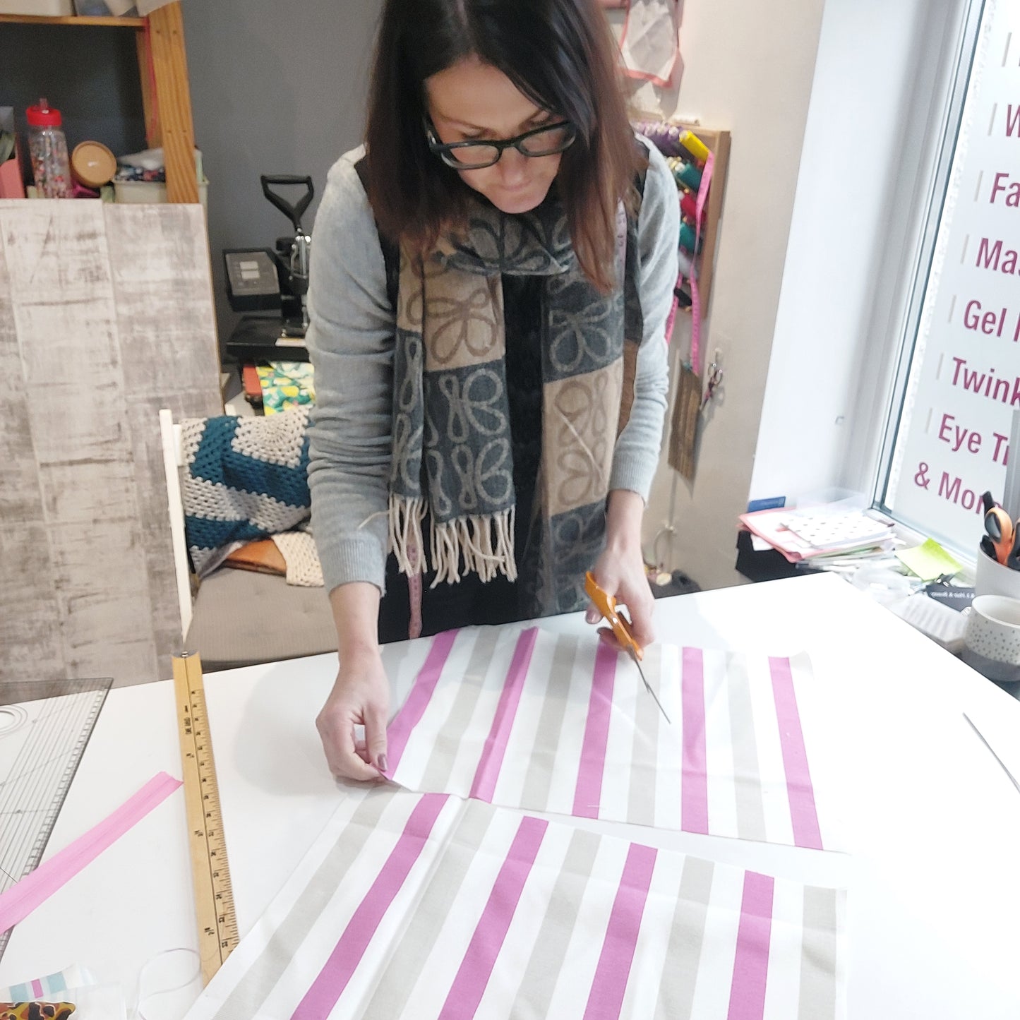 A woman leaning over a work table looking at pieces of fabric with pink, beige and white stripes, holding scissors.