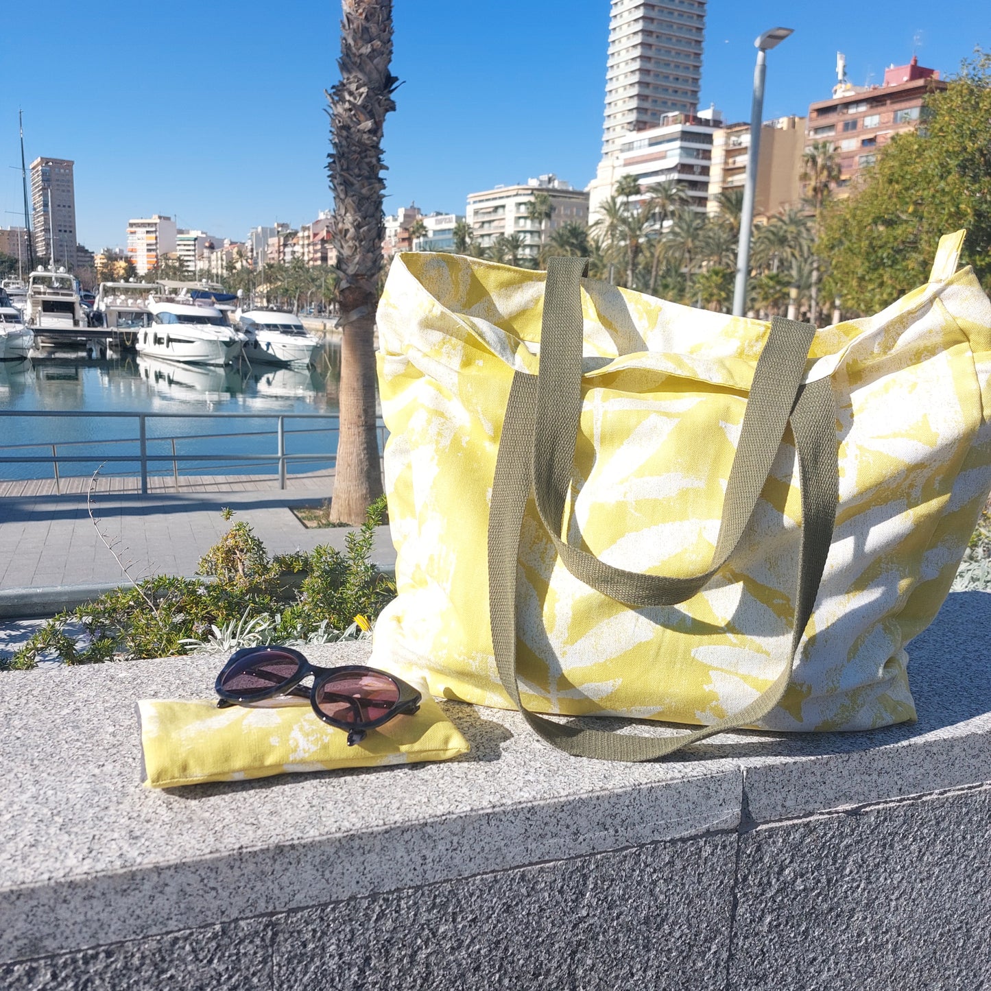 Bright citrus yellow leafy print beach bag pictured with sunglasses and citrus yellow sunglasses case on a sunny harbour.