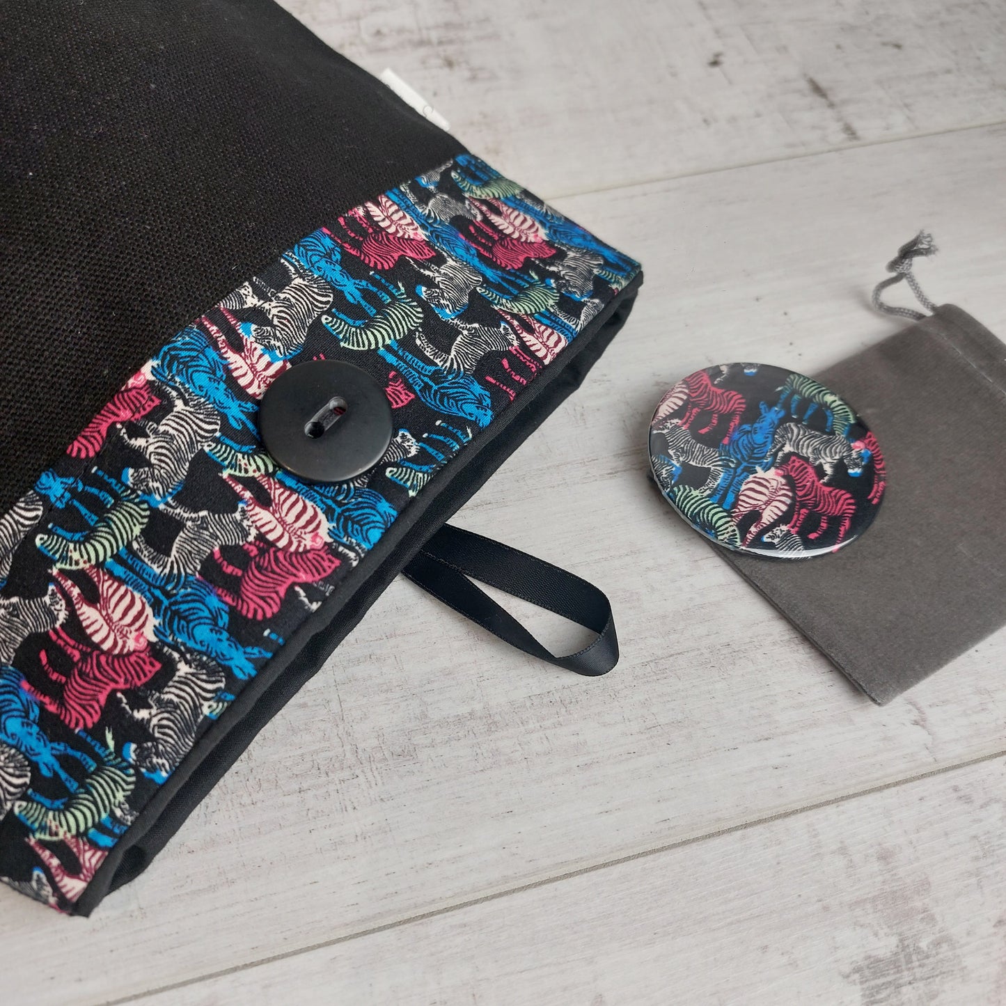 Black button pouch with colourful zebra print top next to a matching zebra print compact mirror on a grey velvet pouch.