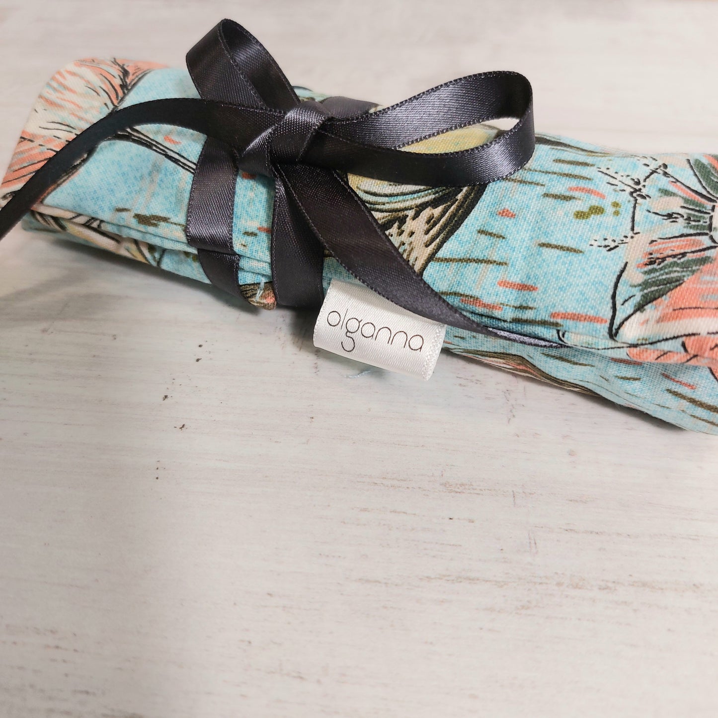 Close up of a rolled up crochet hooks case fastened with grey ribbon and showing olganna branded label. Fabric is sky blue and printed with vanilla and coral.coloured flowers