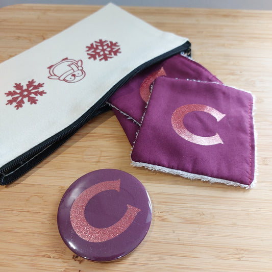 Rich plum glitter initial bamboo face wipes and matching compact mirror with the letter 'C', next to a penguin and snowflake zip pouch.