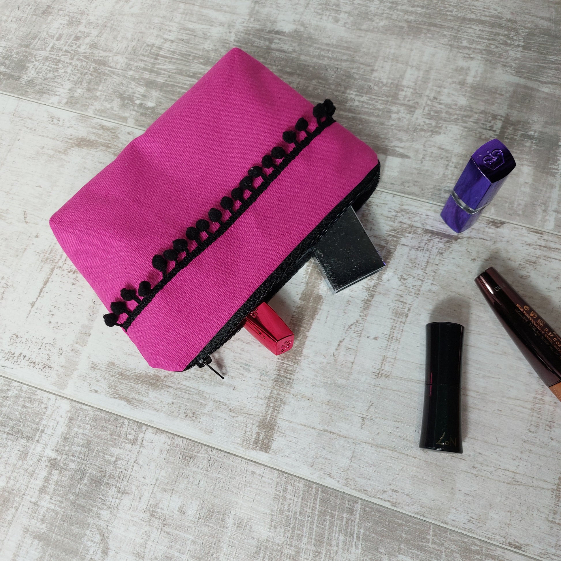 Hot pink canvas makeup bag with black pom poms, pictured with beauty essentials.