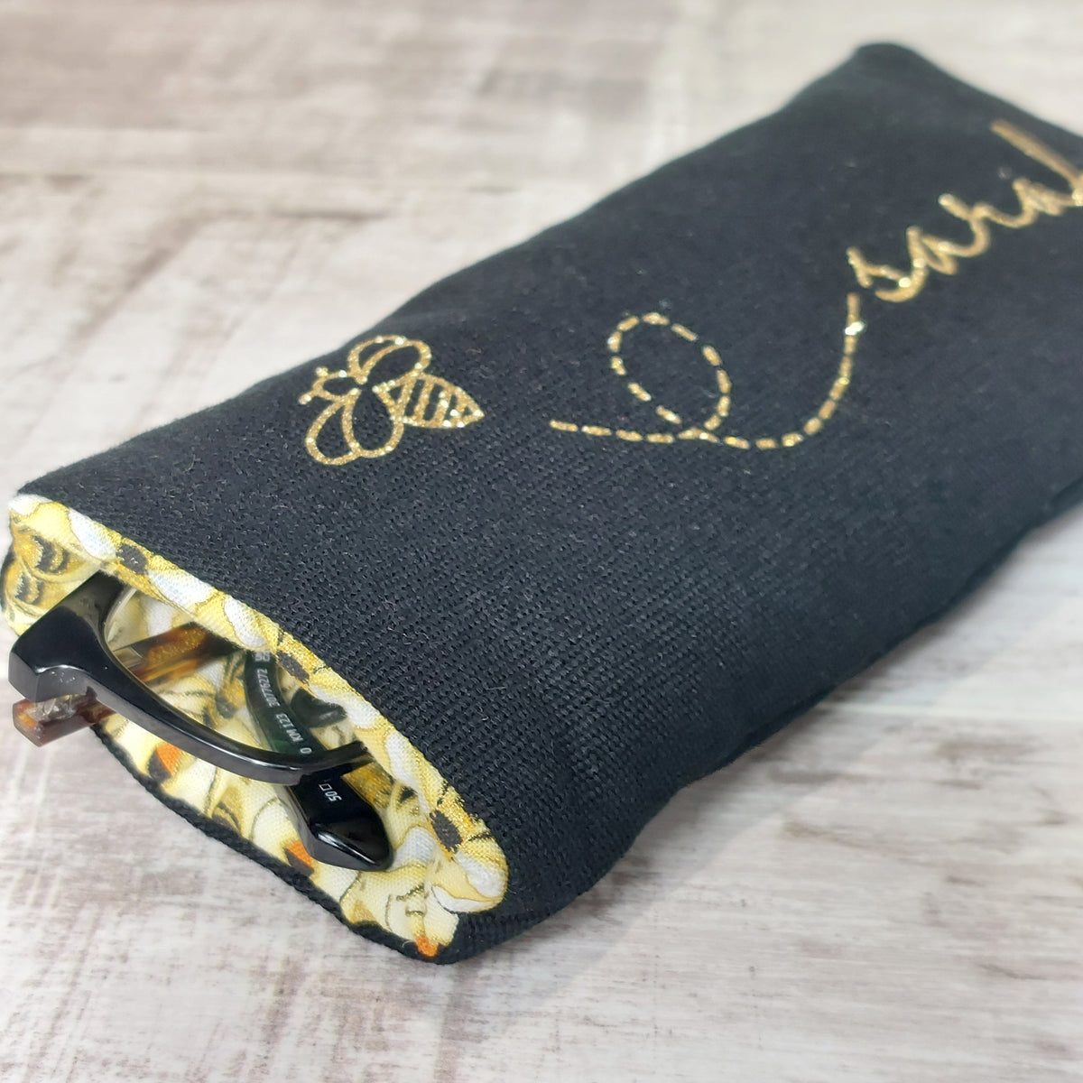 Black soft padded glasses case with gold glitter bee and personalised with the name 'Sarah' in gold, revealing contrast yellow bee lining and glasses inside.