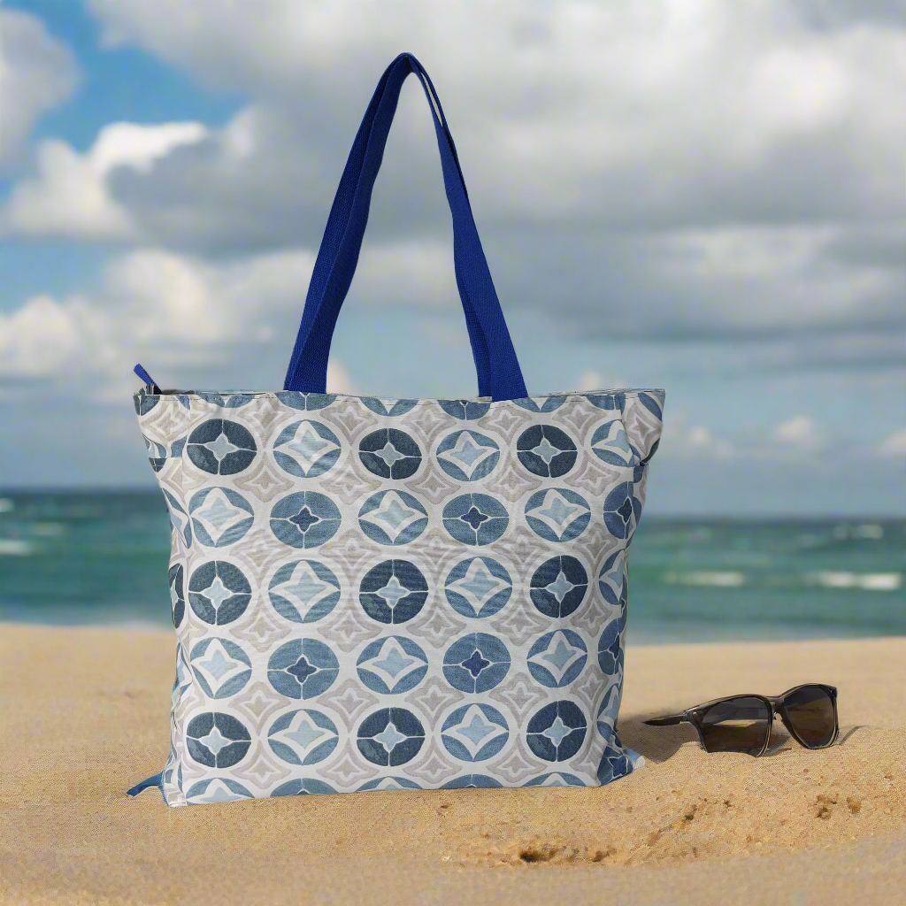 Front view of blue Moroccan tile print beach bag on a beach, pictured with sunglasses.