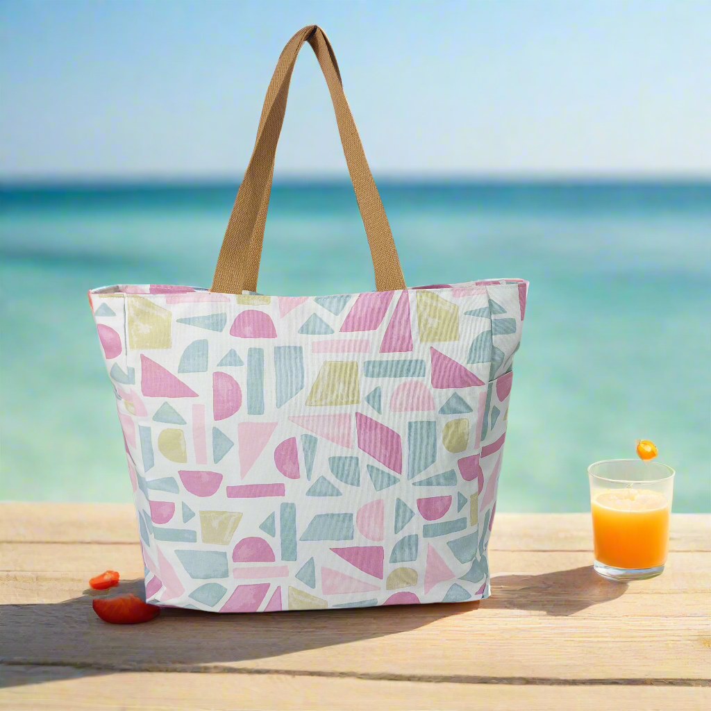 Front view of pink pastels summery beach bag on a picnic bench with ocean backdrop next to a glass of juice.
