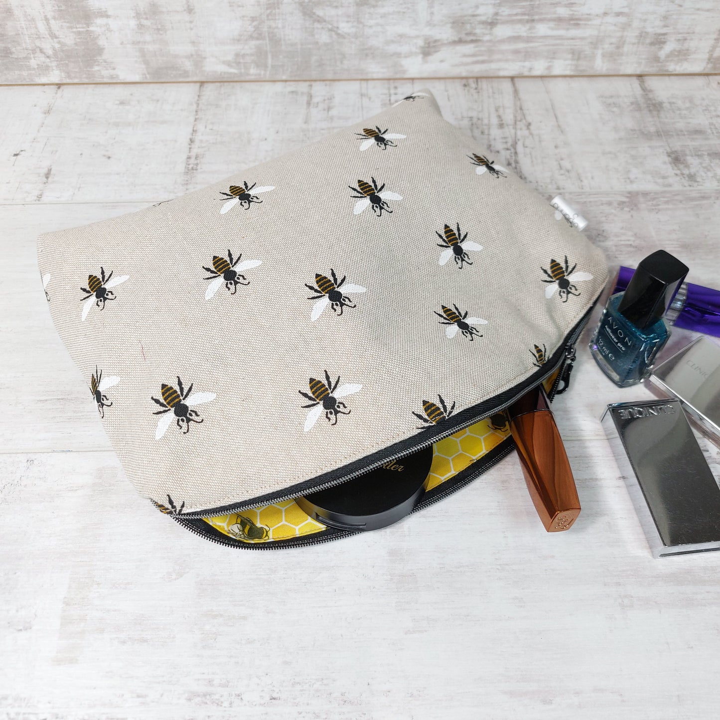 An open makeup bag in repeat bee print on linen-look fabric, revealing a yellow honeycomb pattern lining with makeup items peeking out, next to some nail varnish and makeup items.