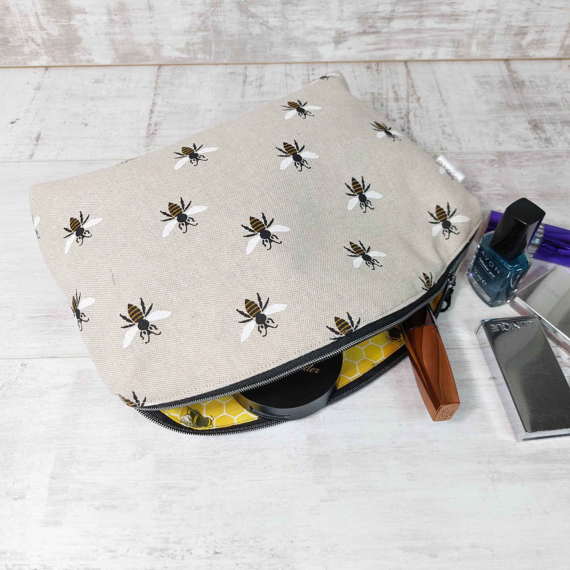 An open makeup bag in repeat bee print on linen-look fabric, revealing a yellow honeycomb pattern lining with makeup items peeking out, next to some nail varnish and makeup items.