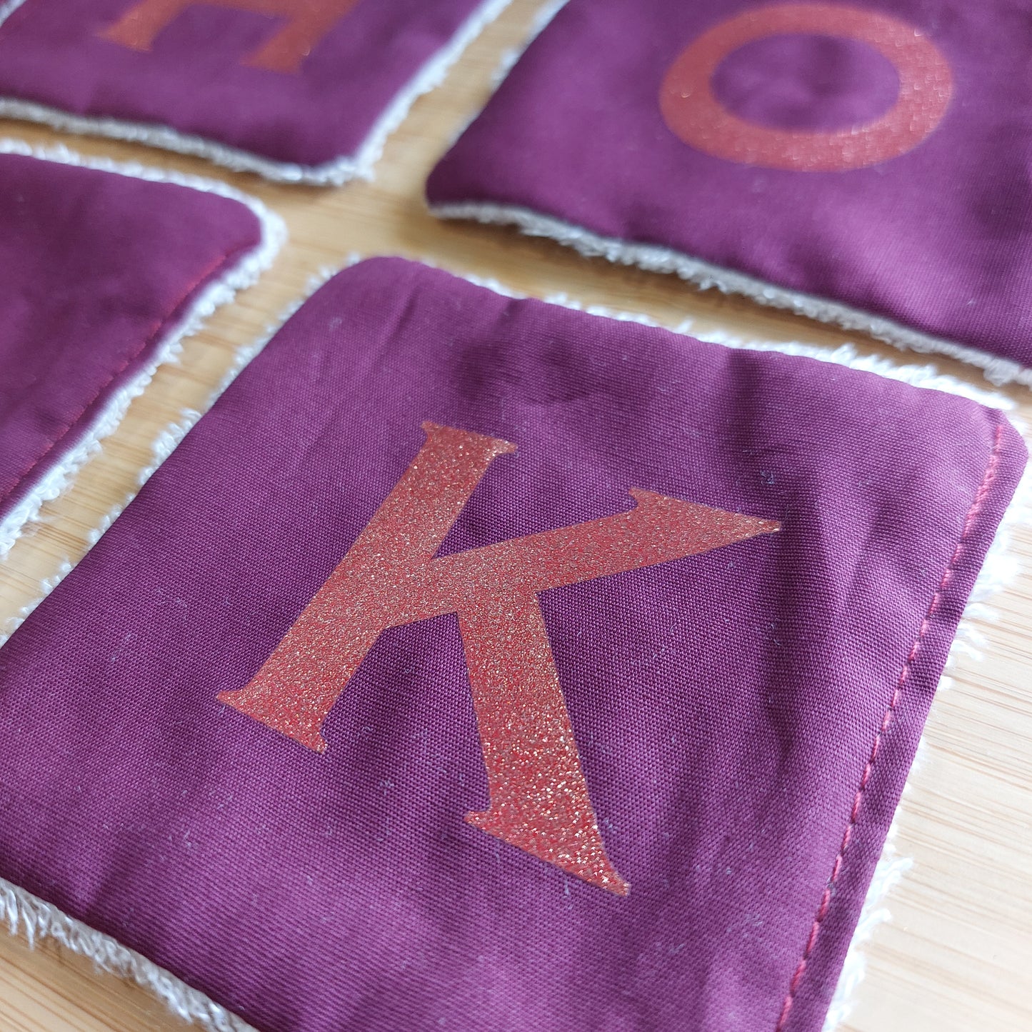 Closeup of four re-usable rich plum face wipes, with different glitter initials, showing a letter 'K' at the front.