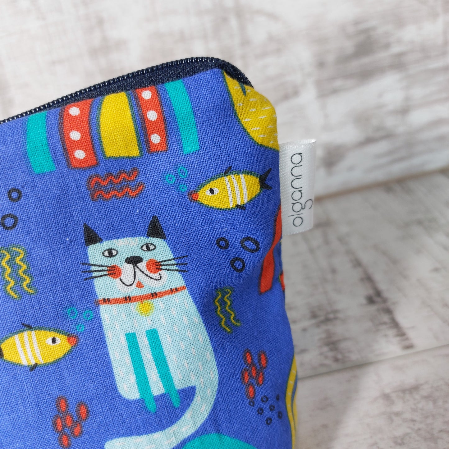 Close-up view of blue rainbow, cat and fish print makeup bag with side label saying 'olganna'.