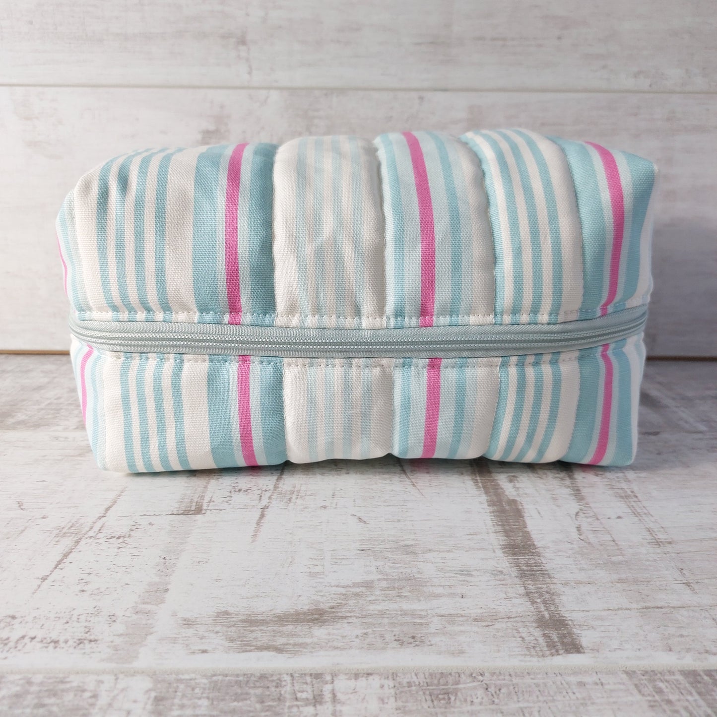 A large quilted boxy toiletries bag in pale blue, pink and white narrow stripes, showing zip fastening.