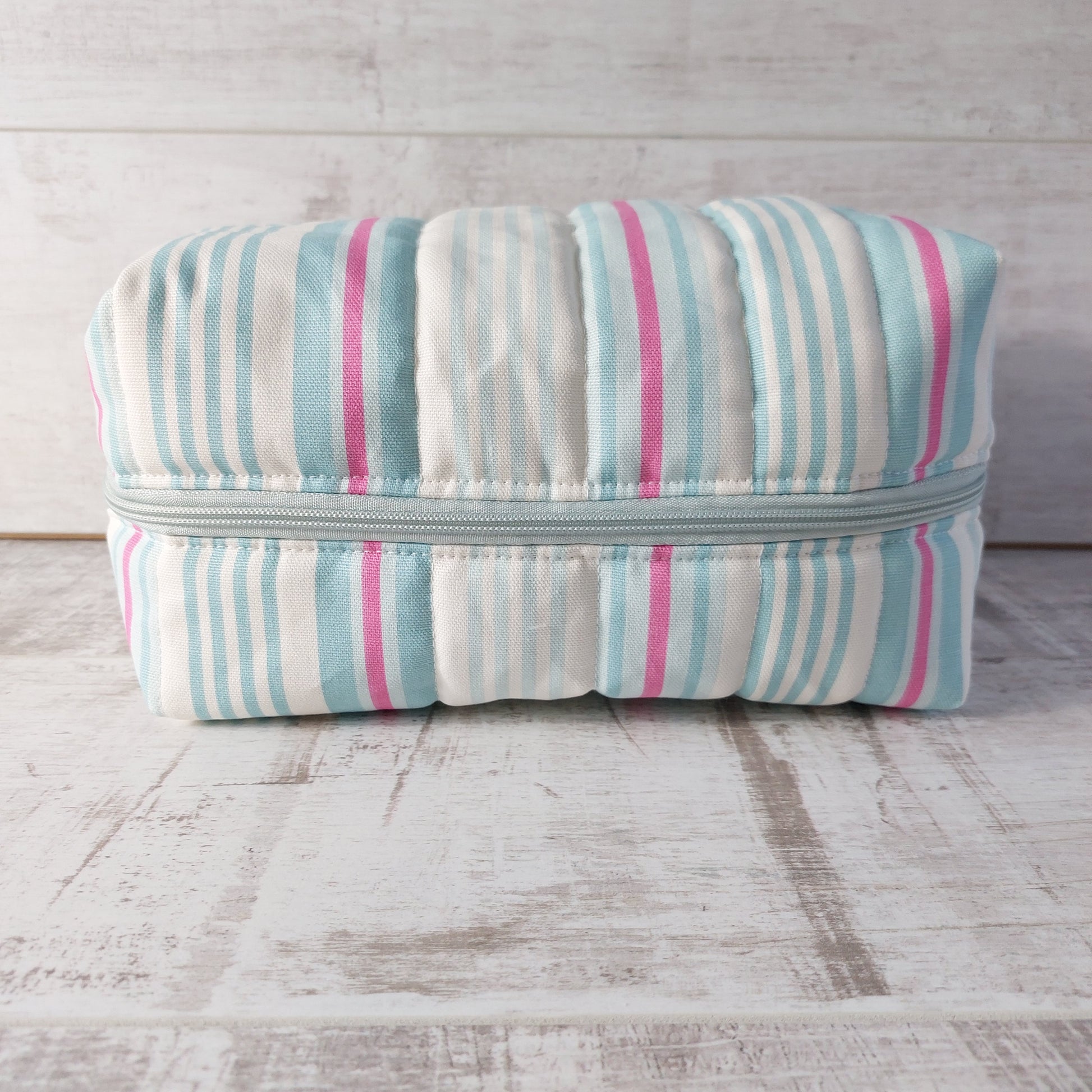 A large quilted boxy toiletries bag in pale blue, pink and white narrow stripes, showing zip fastening.