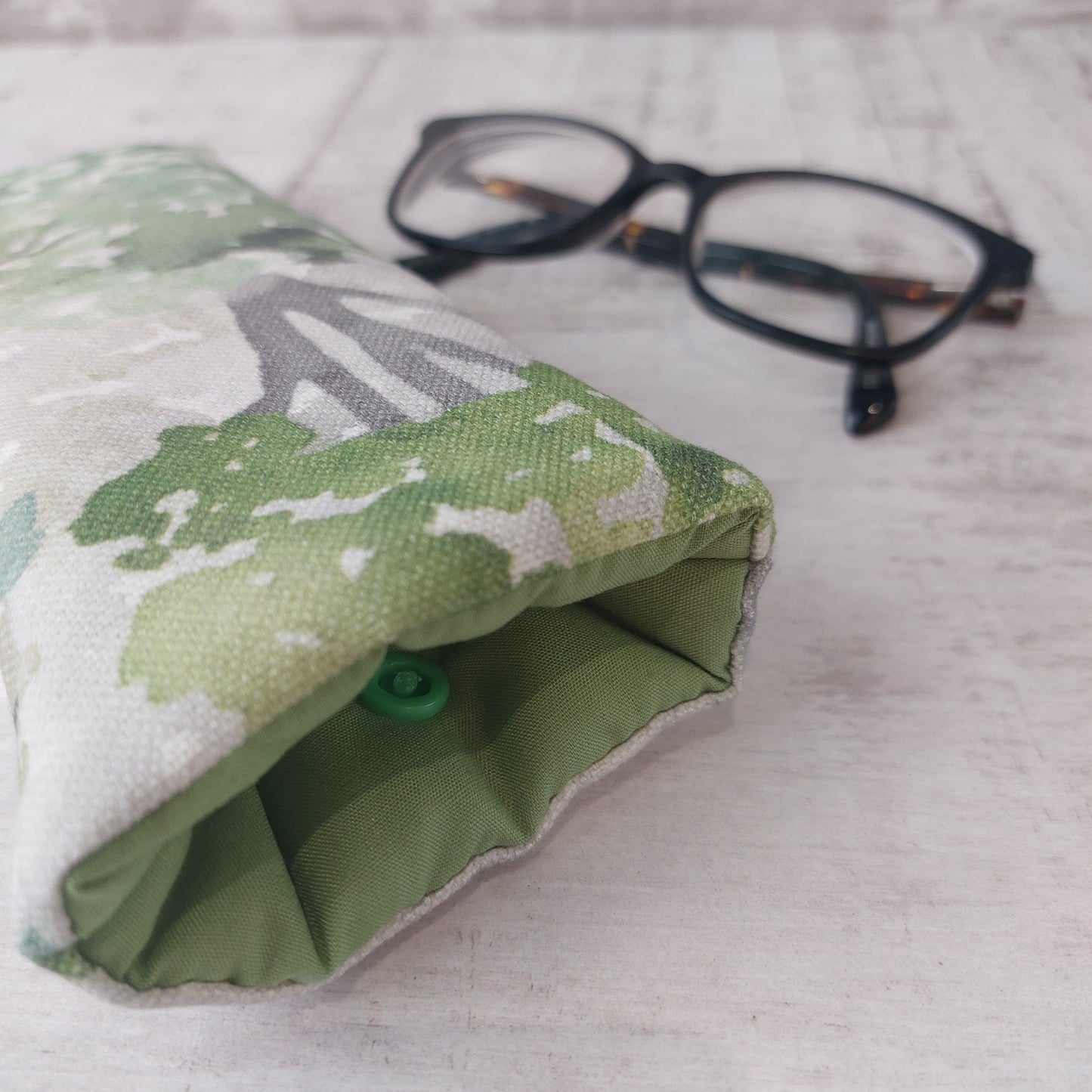 Closeup of green watercolour tree print glasses case with green lining and snap fastening, pictured with glasses.
