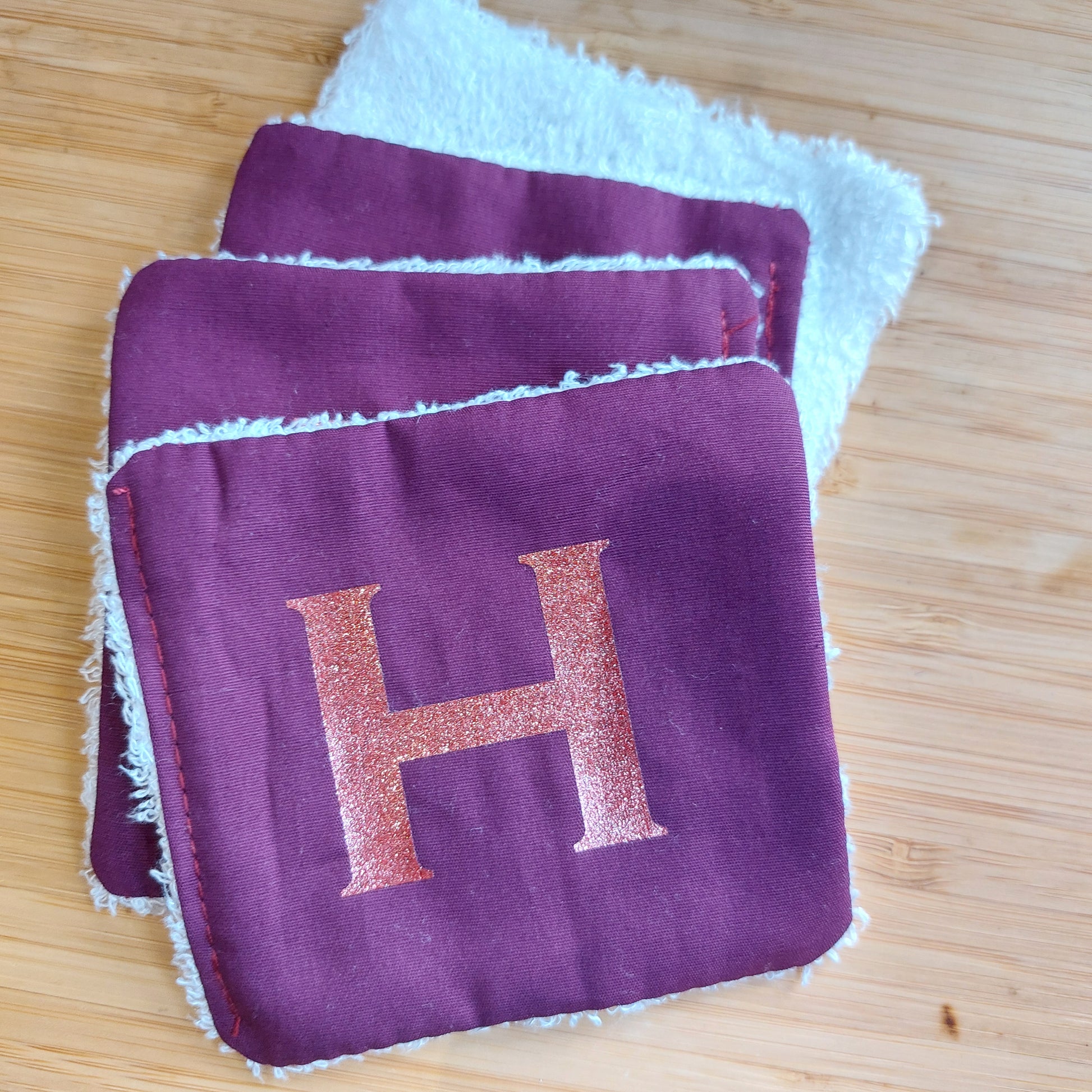 Four re-usable rich plum face wipes, uppermost showing glitter 'H' initial, by Olganna.
