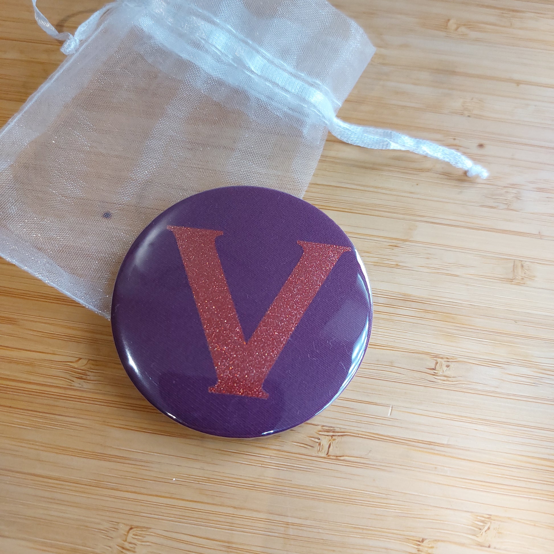 Compact mirror in rich plum with glitter initial 'V' resting on a white organza bag.