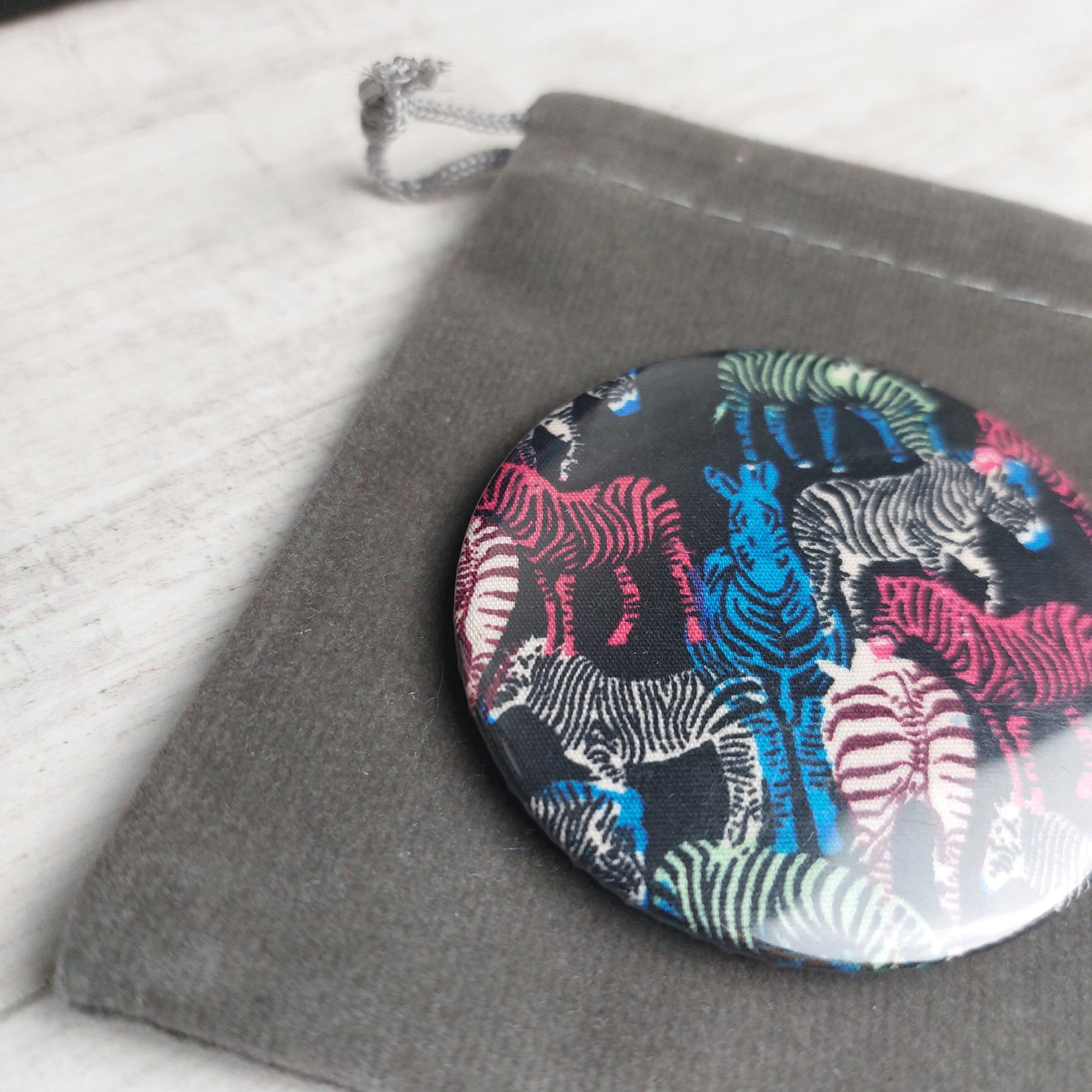 Close up of colourful zebra print compact mirror lying on a grey velvet pouch, by Olganna.