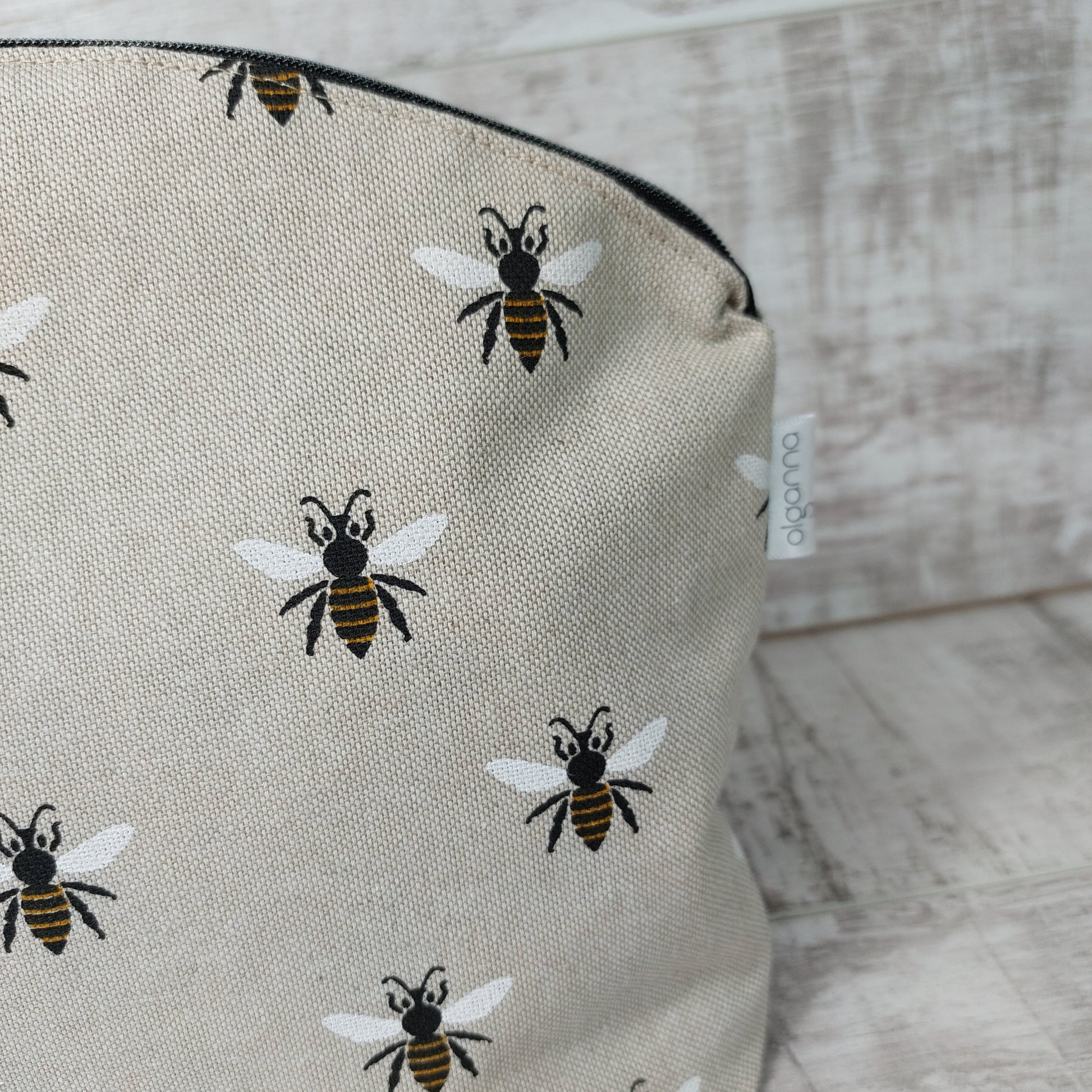 Top corner of a makeup bag standing upright with a zip fastening and repeat bee print on linen-look fabric, displaying a small label stitched in the side seam, saying 'olganna'.