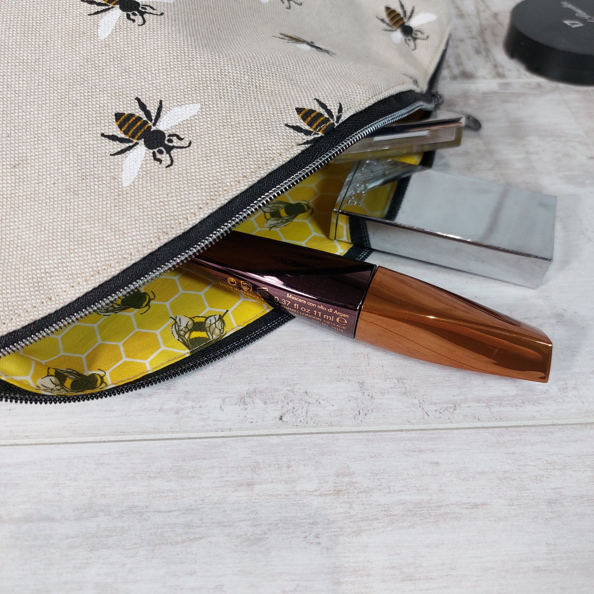 Close up view of an open makeup bag with repeat bee print on linen-look fabric, revealing a yellow honeycomb and bee print lining and with makeup items in the bag partially displayed.