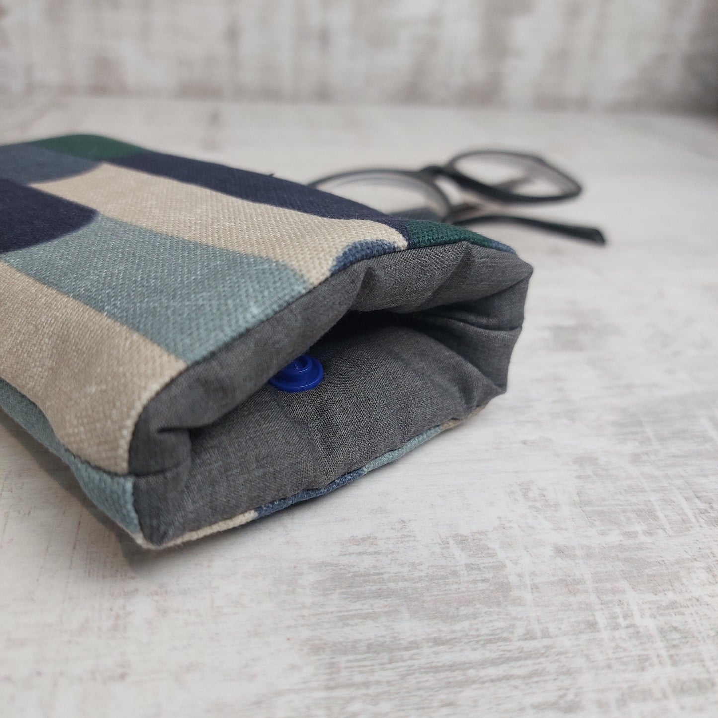 Closeup of a padded glasses case in winter tones showing a grey lining and blue snap fastening with glasses in the background.