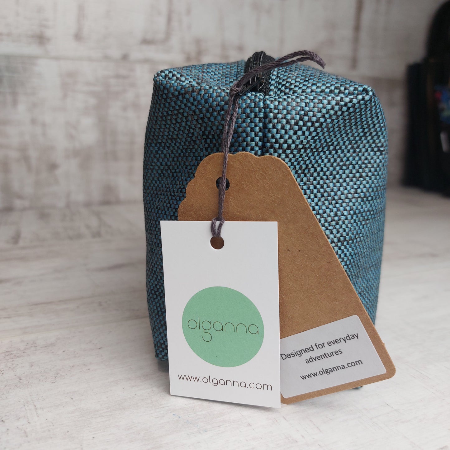 End view of boxy zip pouch in teal and black woven fabric with Olganna gift tags stating 'Designed for everyday adventures www.olganna.com'