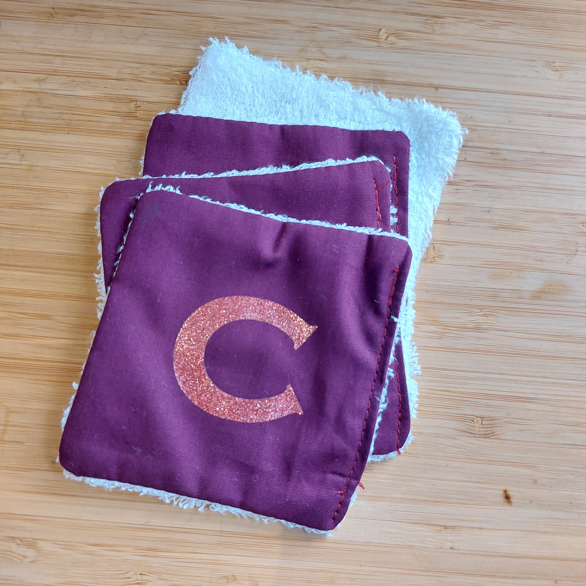 A pile of four re-usable rich plum face wipes, the top one showing glitter 'C' initial and the bottom one with the cotton bamboo facing upwards.