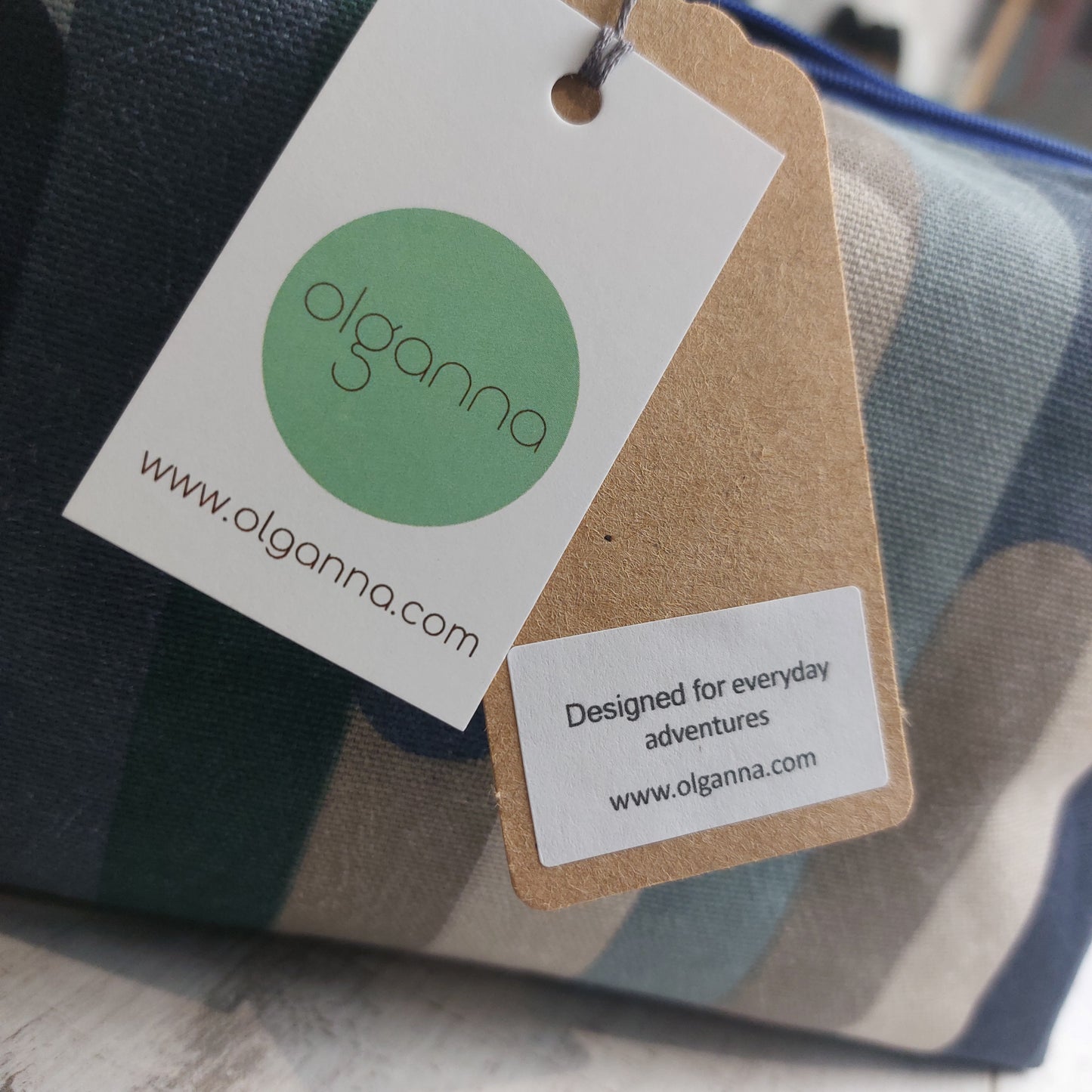 Closeup of Olganna gift tags on a boxy zip pouch in blue and winter tones with a label saying 'Designed for everyday adventures www.olganna.com'