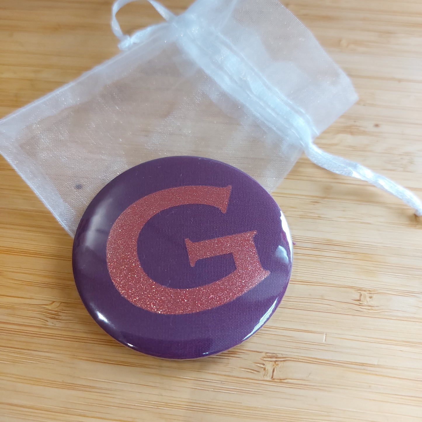 Compact mirror in rich plum with glitter initial 'V' resting on a white organza bag,  by Olganna.