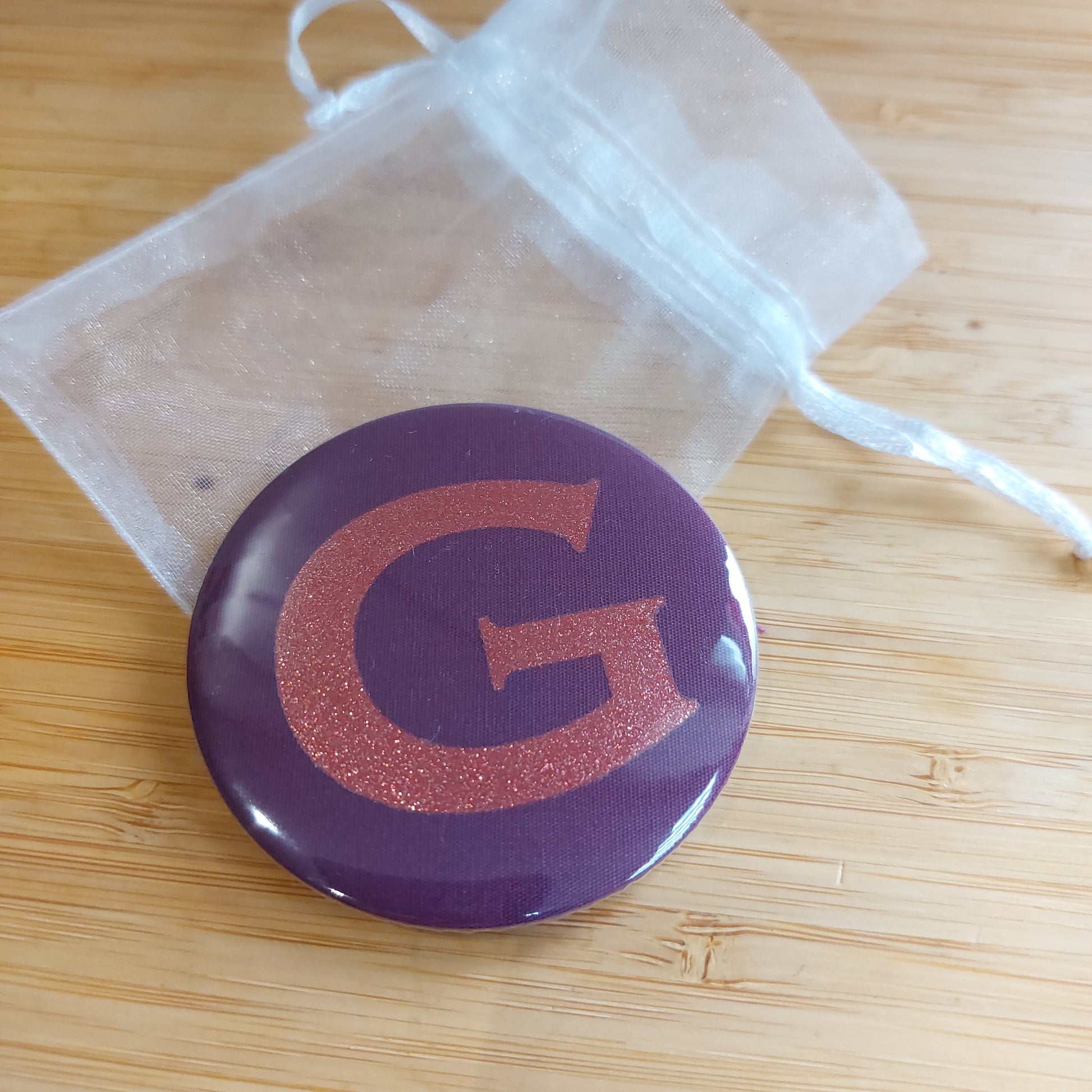 Compact mirror in rich plum with glitter initial 'V' resting on a white organza bag,  by Olganna.