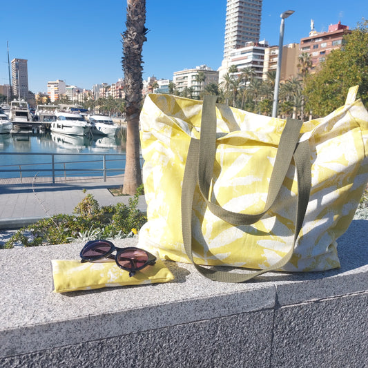Bright citrus yellow leafy print beach bag pictured with sunglasses and citrus yellow sunglasses case on a sunny harbour.