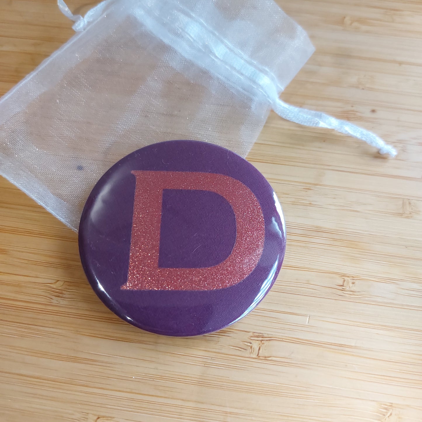 Compact mirror in rich plum with glitter initial 'D' resting on a white organza bag.
