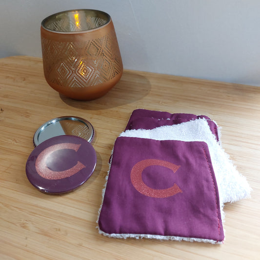 Four re-usable rich plum face wipes, uppermost showing glitter 'C' initial next to two compact mirrors, one displaying rich plum colour reverse and 'C' initial, pictured with a candle.