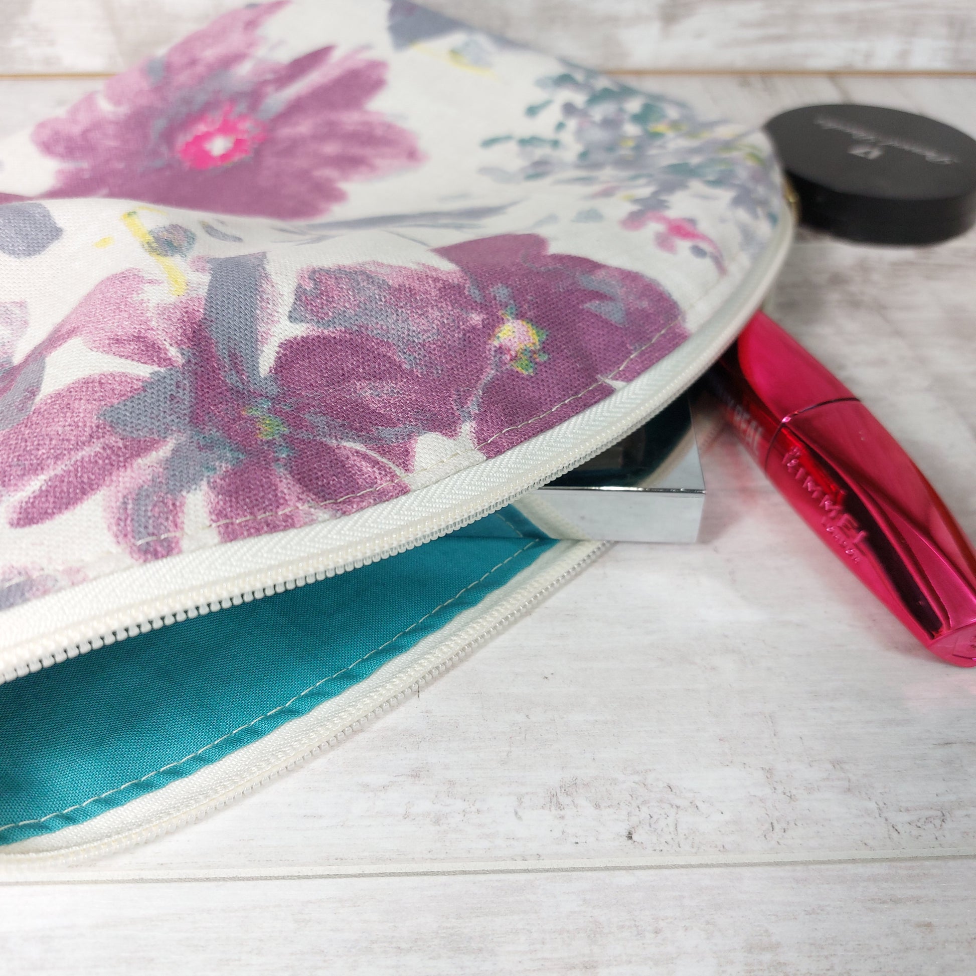 Close up view of the top opening of a watercolour floral makeup bag, unzipped, with teal lining and a mascara wand cascading out.