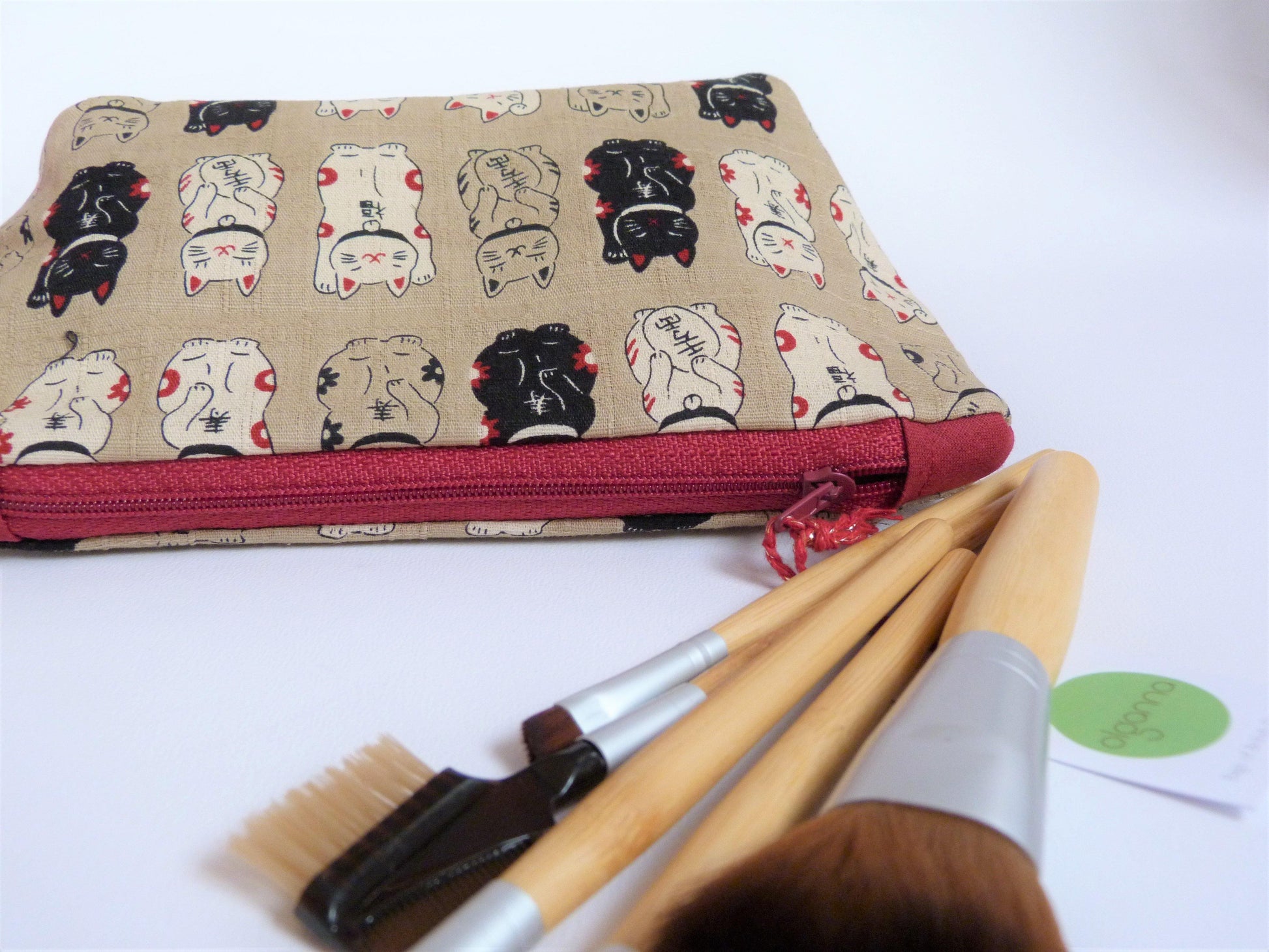 Taupe bag with waving cat print in repeat all over it. Has a red contrast zip and is pictured lying down with wooden handled make up brushes.